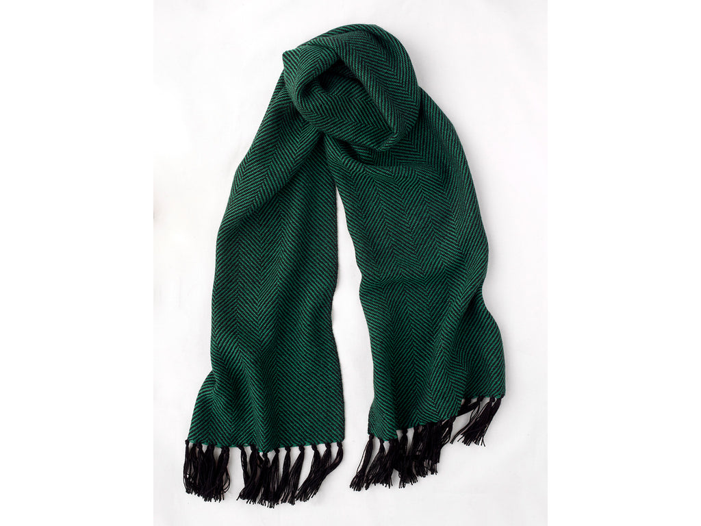 The scarf in the image is a black and dark green alpaca scarf featuring a stylish herringbone pattern. It has black fringing on the ends and comes in a narrow or wide option. It's smooth soft alpaca wool is soft and cosy, making it a perfect accessory for cooler weather.